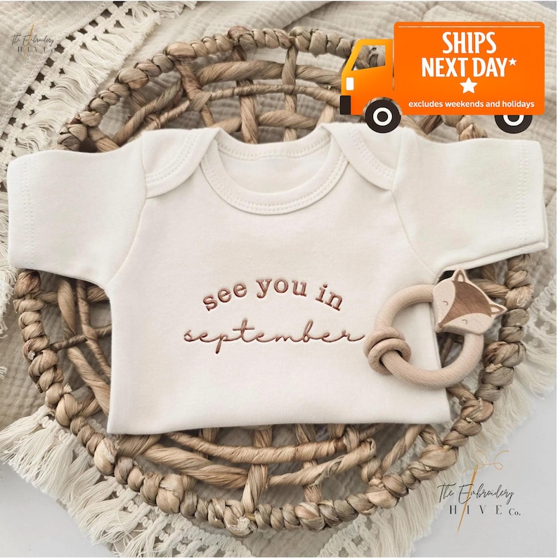 A baby onesie with the text "see you in September" printed on it, placed in a woven basket. There is also a baby rattle next to the onesie.