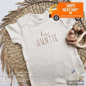 A cream-colored onesie with the text "Hi Auntie" embroidered on it, placed on a woven mat. There is also a toy and a sign that reads "SHIPS NEXT DAY* excludes weekends and holidays".