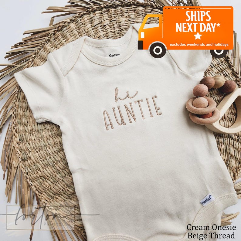 A cream-colored onesie with the text "Hi Auntie" embroidered on it, placed on a woven mat. There is also a toy and a sign that reads "SHIPS NEXT DAY* excludes weekends and holidays".