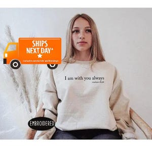 A young woman wearing a white sweatshirt with the text "I am with you always" printed on it, sitting in front of a white background.