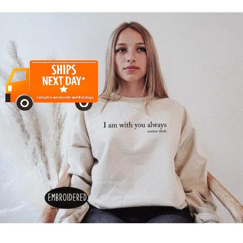 A young woman wearing a white sweatshirt with the text "I am with you always" printed on it, sitting in front of a white background.