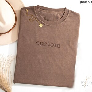 a brown t-shirt with the word "CUSTOM" printed on it, along with a beige hat and some dried flowers.
