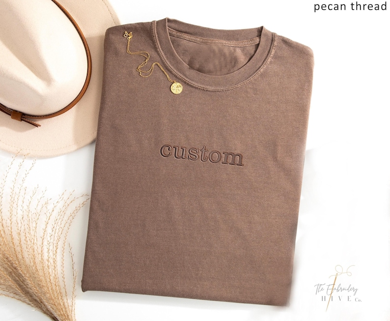 a brown t-shirt with the word "CUSTOM" printed on it, along with a beige hat and some dried flowers.