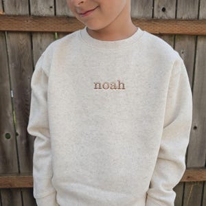 A young boy wearing a cream-colored sweater with the word "noah" embroidered on it.