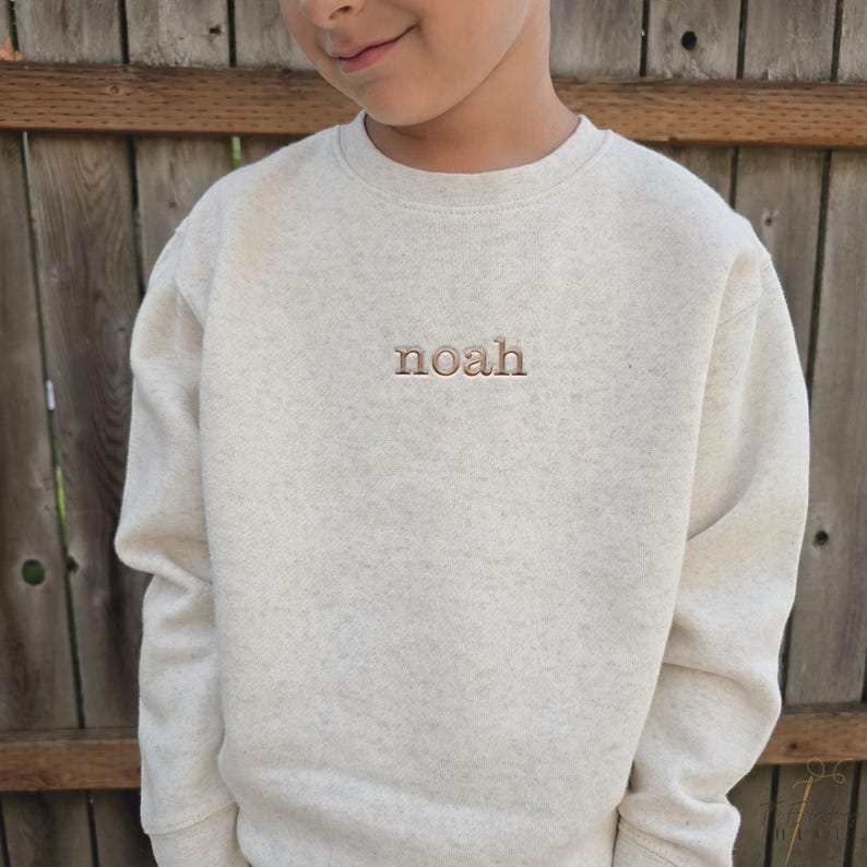 A young boy wearing a cream-colored sweater with the word "noah" embroidered on it.
