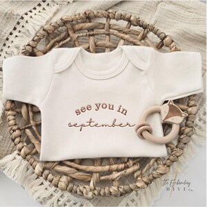 A baby onesie with the text "see you in September" printed on it, placed in a woven basket.