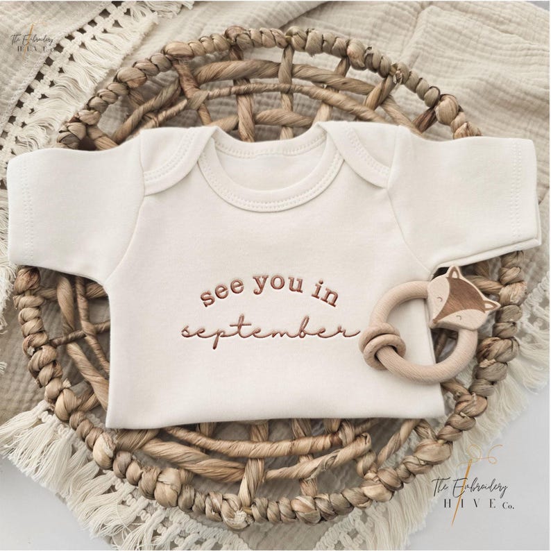 A baby onesie with the text "see you in September" printed on it, placed in a woven basket.