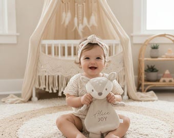 May include: A beige plush deer security blanket featuring embroidered white spots on the head and the name Alice JOY stitched in dark thread on the front. The soft fabric toy has a rounded body, small ears, and a pointed nose, held by a smiling child.