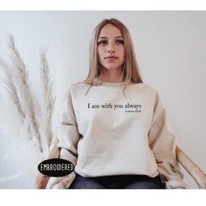 A young woman wearing a beige sweatshirt with the text "I am with you always" printed on it, sitting in a chair against a plain background.