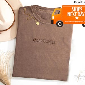 a brown t-shirt with the word "CUSTOM" printed on it, along with a hat and a beige ribbon. There is also a text overlay that reads "SHIPS NEXT DAY" and "excluses weekends and holidays".
