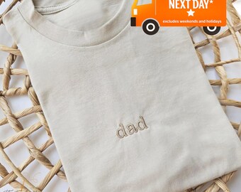 Puede incluir: Una camiseta beige con la palabra "dad" bordada en hilo a juego. La camiseta está doblada sobre una superficie de mimbre tejido. La imagen incluye un gráfico de un camión de reparto.