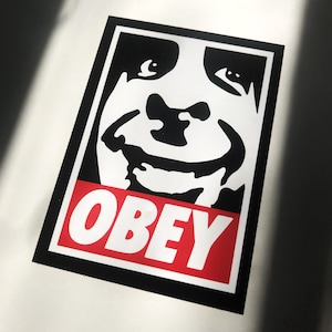 Op de afbeelding: Een rechthoekige sticker met een gestileerd zwart-wit gezicht en het woord "OBEY" in witte letters op een rode achtergrond. De sticker heeft een zwarte rand en een witte achtergrond.