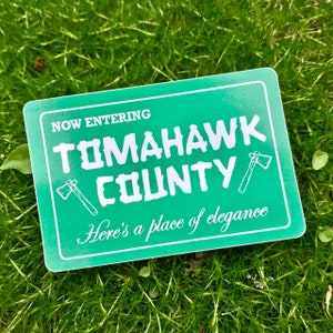Op de afbeelding: Een rechthoekige sticker met een groene achtergrond en witte rand. De tekst "NOW ENTERING TOMAHAWK COUNTY" wordt weergegeven, samen met twee illustraties van bijlen. De zin "Here's a place of elegance" is ook opgenomen.