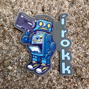 Op de afbeelding: Een blauwe robot met gele ogen en koptelefoon die een cassettebandje vasthoudt. De robot draagt sneakers en heeft een grijze achtergrond met het woord "ÍROKK" in blauwe letters.