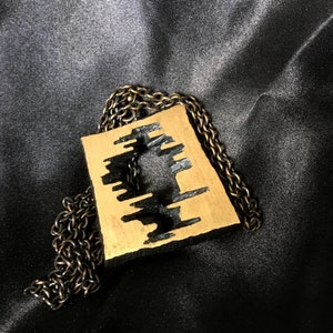 May include: A gold-colored pendant with a black, abstract design, suspended from a dark chain. The pendant has a rectangular shape with a cut-out pattern. The chain and pendant rest on a black, silky fabric.