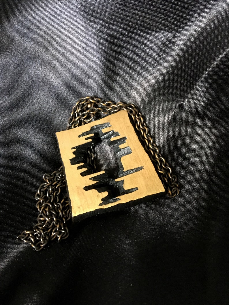 May include: A gold-colored pendant with a black, abstract design, suspended from a dark chain. The pendant has a rectangular shape with a cut-out pattern. The chain and pendant rest on a black, silky fabric.