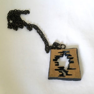 May include: A unique necklace featuring a rectangular pendant with a cut-out design. The pendant has a tan center with black accents and a dark chain. The necklace is a statement piece, perfect for adding a touch of artistry to any outfit.