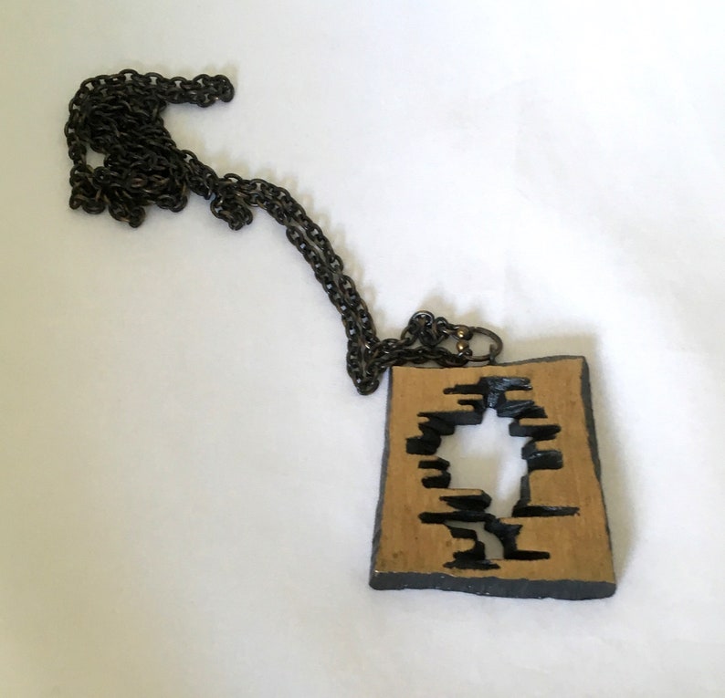 May include: A unique necklace featuring a rectangular pendant with a cut-out design. The pendant has a tan center with black accents and a dark chain. The necklace is a statement piece, perfect for adding a touch of artistry to any outfit.