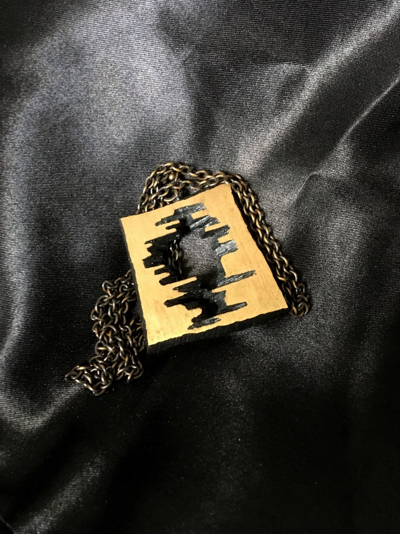 May include: A gold-colored pendant with a geometric design and black accents hangs from a dark chain. The pendant has a rectangular shape with a cut-out pattern. The necklace is displayed on a dark, textured surface.