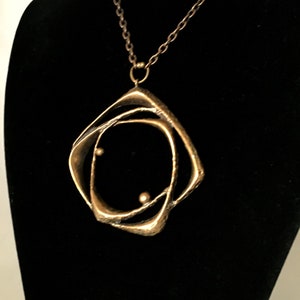 May include: A gold-coloured pendant necklace with an abstract design. The pendant has an open, geometric shape with two small, spherical accents. The necklace hangs on a delicate chain against a black backdrop.