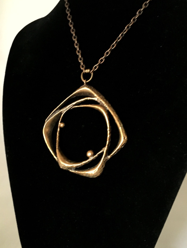 May include: A gold-coloured pendant necklace with an abstract design. The pendant has an open, geometric shape with two small, spherical accents. The necklace hangs on a delicate chain against a black backdrop.