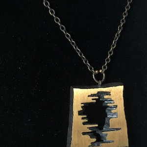 May include: A unique necklace featuring a rectangular pendant with a gold-colored center and a black, textured border. The pendant has a cut-out design. The necklace has a double-strand, antique-bronze chain.