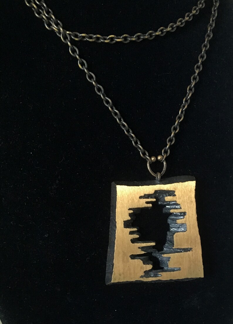 May include: A unique necklace featuring a rectangular pendant with a gold-colored center and a black, textured border. The pendant has a cut-out design. The necklace has a double-strand, antique-bronze chain.