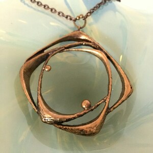 May include: A bronze pendant necklace with an abstract design. The pendant features a series of curved, interconnected shapes, including a central circle and a square-like outer frame. The necklace has a delicate chain.