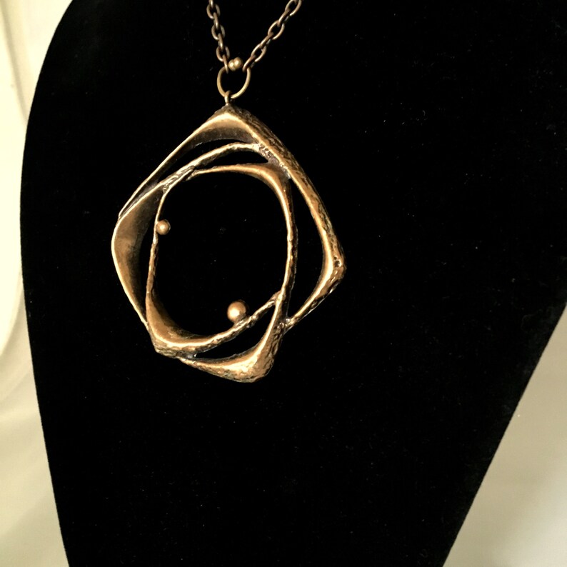 May include: A bronze pendant necklace with an abstract, geometric design. The pendant has a layered, openwork structure with two small, spherical accents. The necklace hangs on a delicate chain, displayed against a black background.