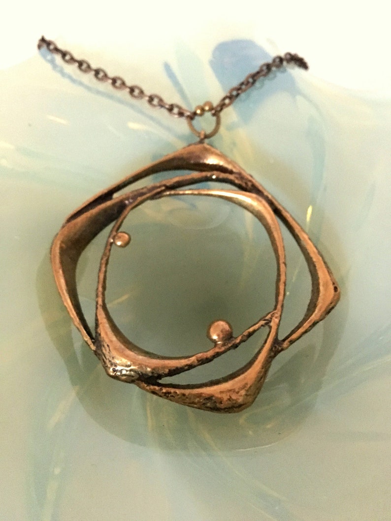 May include: A bronze pendant necklace with an abstract design. The pendant has a central circular element surrounded by curved, open shapes. The necklace has a delicate chain and a small clasp. The pendant is approximately 5 cm in diameter.