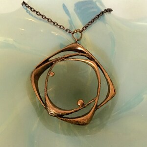 May include: A bronze pendant necklace with an abstract design. The pendant has a central circle and an outer, angular shape, connected by curved lines. The chain is a dark, linked metal. The necklace is displayed on a light blue surface.