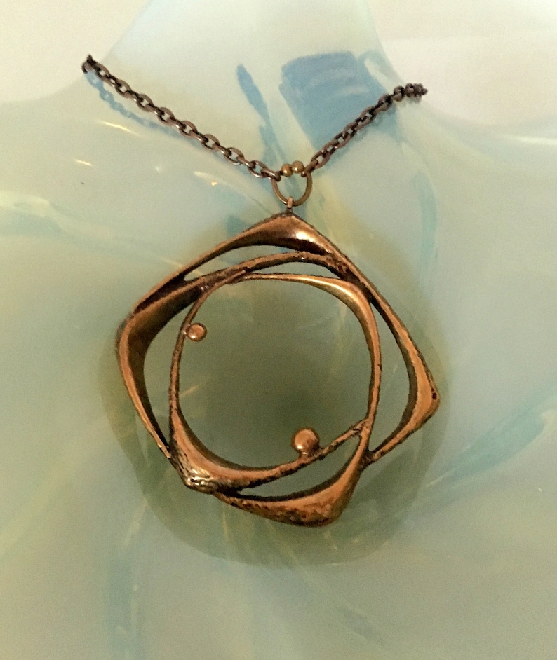 May include: A bronze pendant necklace with an abstract design. The pendant has a central circle and an outer, angular shape, connected by curved lines. The chain is a dark, linked metal. The necklace is displayed on a light blue surface.