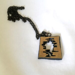 May include: A square pendant necklace with a dark chain. The pendant has a gold-colored center with a black, abstract design cut out. The edges of the pendant are black. The necklace is displayed on a white surface.