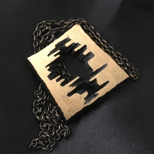 May include: A square pendant necklace with a gold-colored surface and a black, abstract design. The pendant is suspended from a dark, chain necklace. The necklace is displayed on a black background.