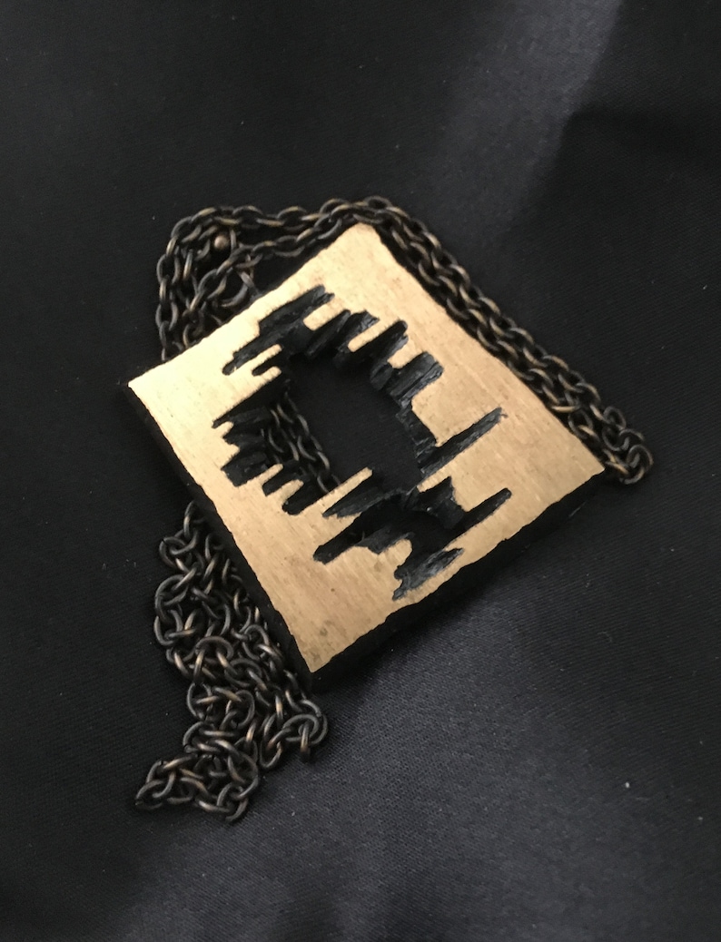 May include: A square pendant necklace with a gold-colored surface and a black, abstract design. The pendant is suspended from a dark, chain necklace. The necklace is displayed on a black background.