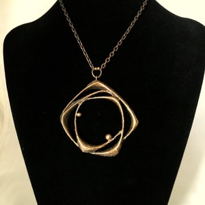 May include: A gold-coloured pendant necklace with a geometric design. The pendant has a square frame with an abstract, swirling interior and two small, spherical accents. The chain is a dark, antique bronze colour, and the necklace is displayed on a black mannequin.