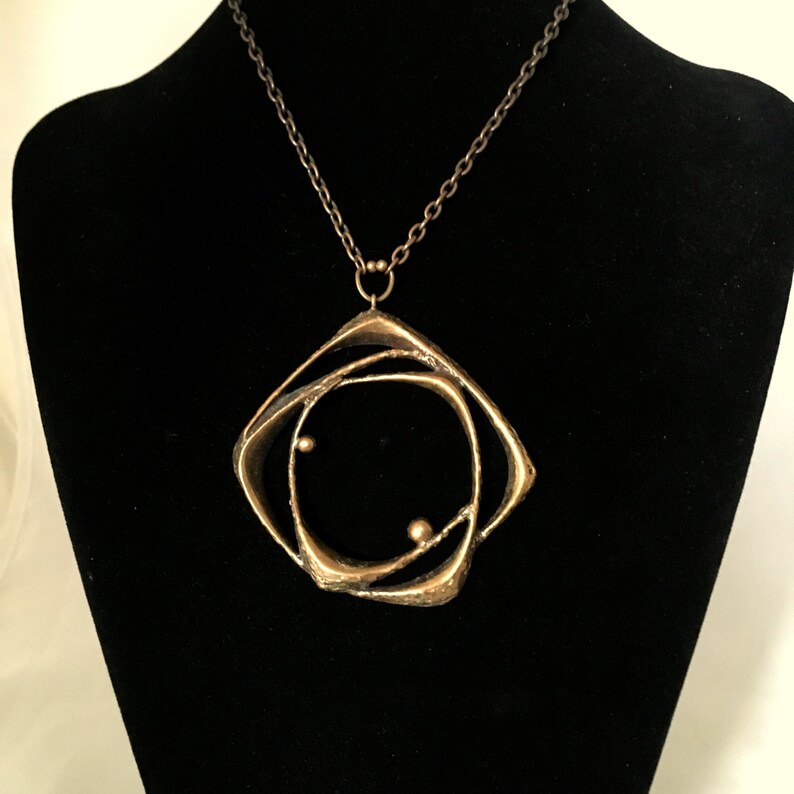 May include: A gold-coloured pendant necklace with a geometric design. The pendant has a square frame with an abstract, swirling interior and two small, spherical accents. The chain is a dark, antique bronze colour, and the necklace is displayed on a black mannequin.