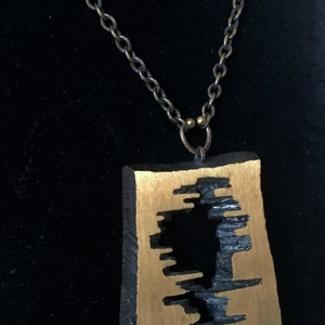 May include: A unique pendant necklace featuring a rectangular, gold-toned pendant with a black, abstract design. The pendant hangs from a dark, linked chain. The necklace is displayed against a black background.