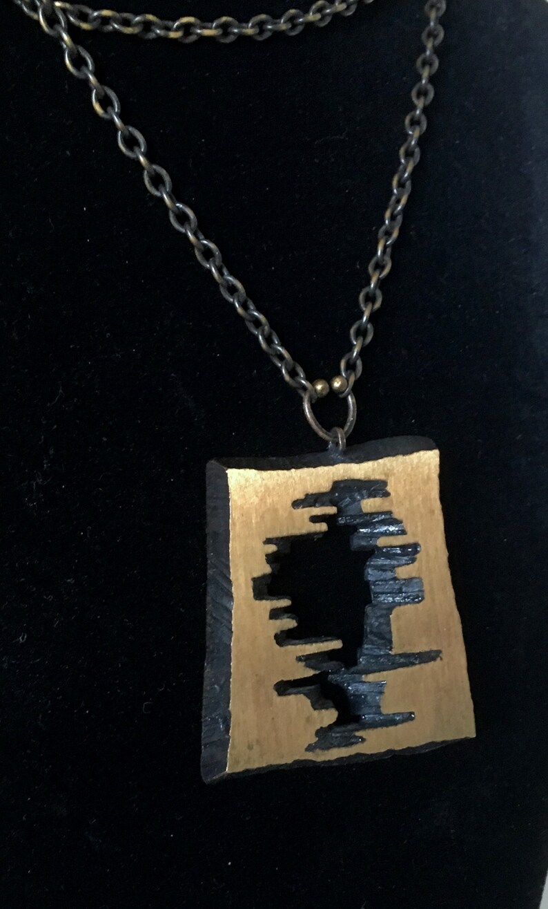 May include: A unique pendant necklace featuring a rectangular, gold-toned pendant with a black, abstract design. The pendant hangs from a dark, linked chain. The necklace is displayed against a black background.