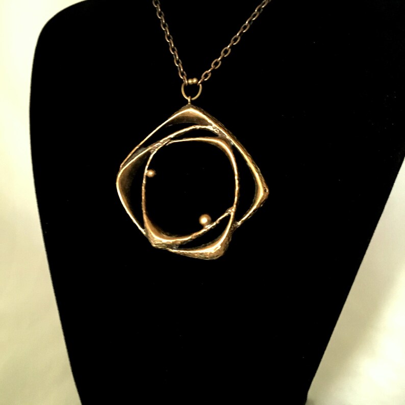 May include: A gold-coloured pendant necklace with an abstract, geometric design. The pendant has a square frame with curved interior lines and two small, spherical accents. The necklace hangs on a delicate chain against a black backdrop.