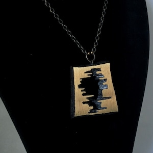 May include: A handcrafted pendant necklace featuring a rectangular, gold-toned pendant with a black, abstract design. The pendant hangs from a layered, dark chain. The necklace is displayed on a black bust.