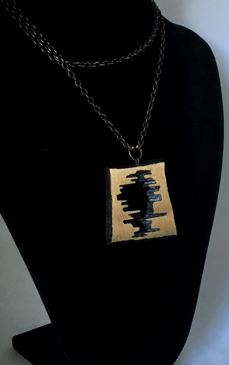 May include: A handcrafted pendant necklace featuring a rectangular, gold-toned pendant with a black, abstract design. The pendant hangs from a layered, dark chain. The necklace is displayed on a black bust.