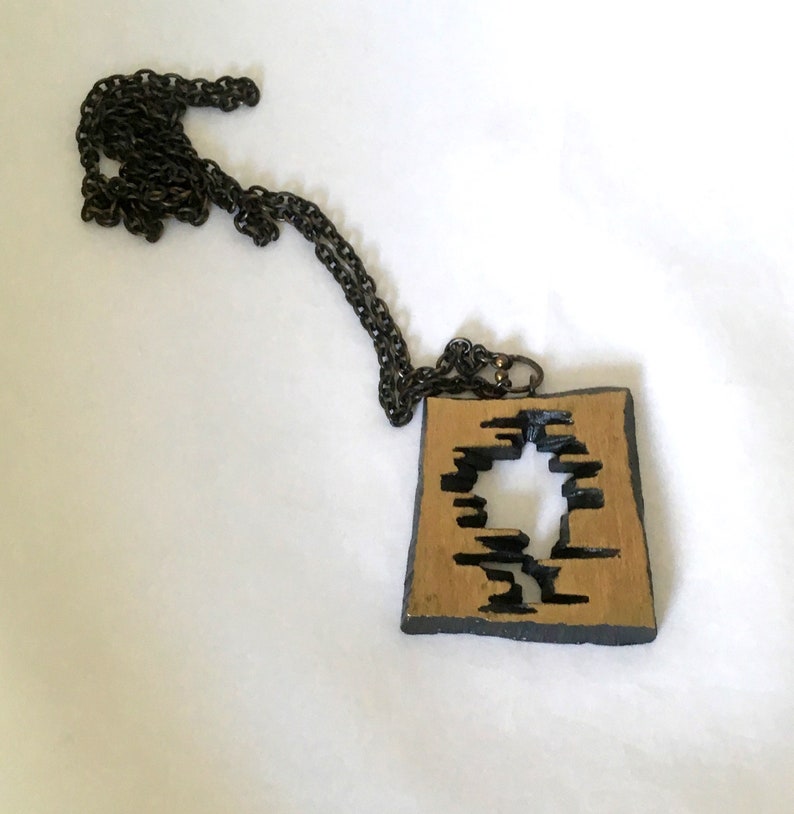 May include: A square pendant necklace with a dark chain. The pendant has a gold-colored center with a black, abstract design cut out. The edges of the pendant are black.