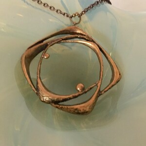 May include: A bronze pendant necklace with an abstract geometric design. The pendant features a central circle within a square-like frame, with small spherical accents. The necklace has a delicate chain.
