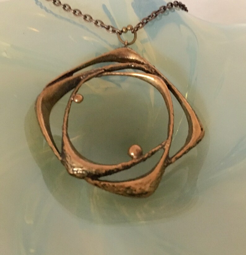 May include: A bronze pendant necklace with an abstract geometric design. The pendant features a central circle within a square-like frame, with small spherical accents. The necklace has a delicate chain.