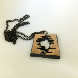 May include: A wooden pendant necklace with a dark chain. The pendant is square with a natural wood color and a black, abstract design cut out of the center. The edges of the pendant are also black.