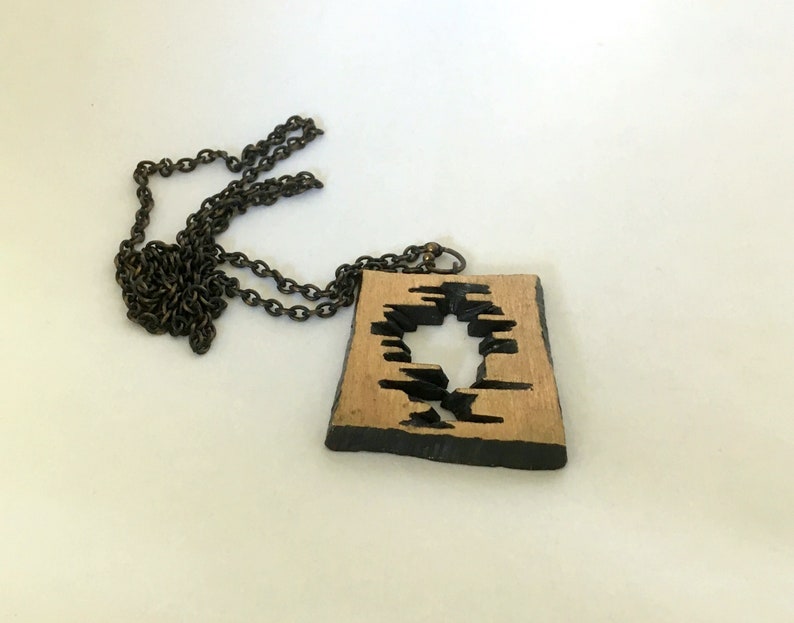May include: A wooden pendant necklace with a dark chain. The pendant is square with a natural wood color and a black, abstract design cut out of the center. The edges of the pendant are also black.