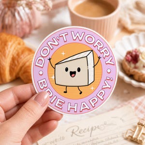 Può includere: Un adesivo circolare raffigurante una fetta di formaggio brie in stile cartoon con un volto sorridente, braccia e gambe. Lo sfondo è arancione con stelline bianche, circondato da un bordo rosa con il testo DON'T WORRY BRIE HAPPY in bianco.