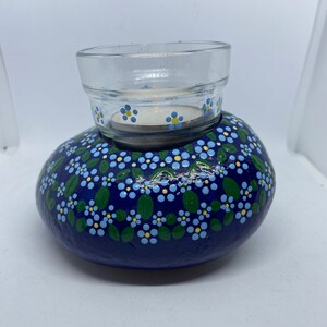 Puede incluir: Una roca pintada de azul con un dise&ntilde;o floral blanco y amarillo, que sostiene un portavelas de vidrio.