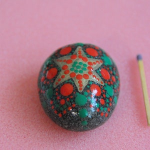 May include: A hand-painted rock with a green and red star design. The rock is about the size of a matchstick.